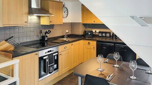 Cocina compacta con armarios de madera, horno empotrado, lavamanos y comedor con sillas y copas de vino.