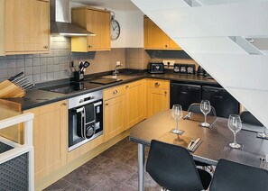 Een compacte keuken met houten kastjes, een inbouwoven, een spoelbak en een eethoek met stoelen en wijnglazen.