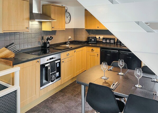 Et lille køkken med træskabe, indbygget ovn, vask og spiseplads med stole og vinglas.