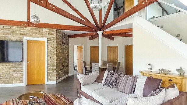 Amplia sala de estar con techo alto, vigas de madera a la vista, pared de ladrillo, televisor de pantalla plana, sofá con cojines estampados y una escalera que conduce a un nivel superior.
