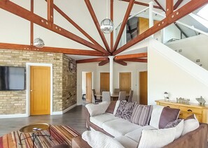 Amplia sala de estar con techo alto, vigas de madera a la vista, pared de ladrillo, televisor de pantalla plana, sofá con cojines estampados y una escalera que conduce a un nivel superior.