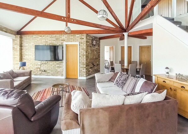 Amplia sala de estar con techo alto, vigas de madera a la vista, chimenea de piedra, televisor de pantalla plana y comedor con mobiliario moderno.