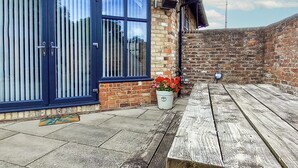 Un patio con un banco de madera, una planta en maceta con flores rojas y un muro de ladrillo.