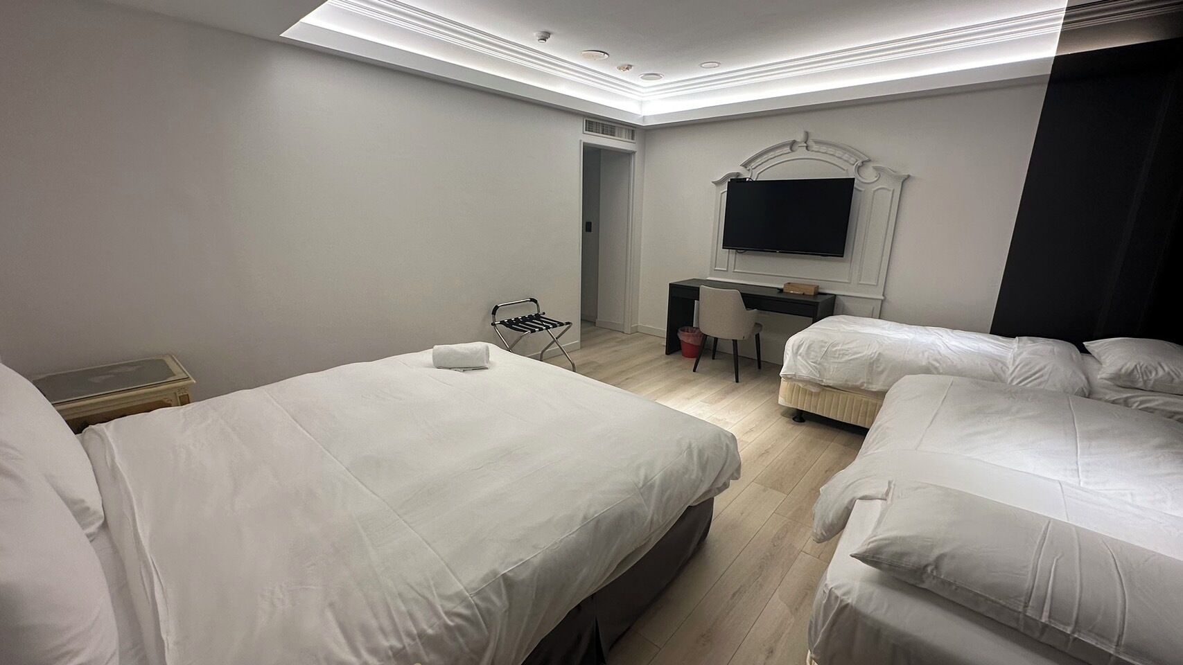 A hotel room with two single beds, a desk with a chair, a flat-screen TV mounted on the wall, and a small seating area with a chair and a table.