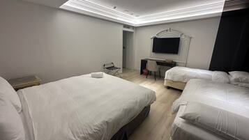 A hotel room with two single beds, a desk with a chair, a flat-screen TV mounted on the wall, and a small seating area with a chair and a table.