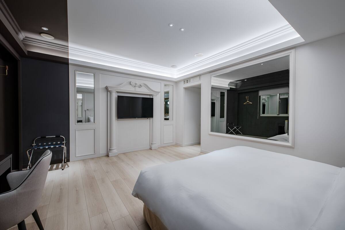 Un dormitorio moderno con una cama grande, un televisor en la pared, un espejo y una zona de estar con una silla y una mesita.