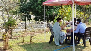 A group of people seated at an outdoor table under a red canopy, surrounded by greenery and a river. - The Raajas - Camp & Resorts (Yamkeshwar)