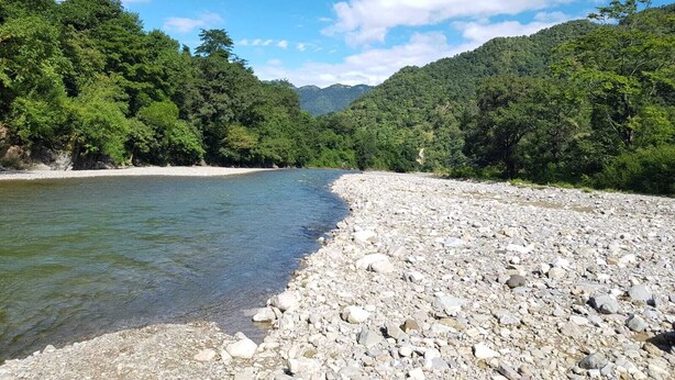 A riverbank with a rocky shoreline and dense forest on both sides. - The Raajas - Camp & Resorts (Yamkeshwar)
