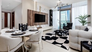 A modern living room with a dining area, a flat-screen TV mounted on a wooden panel, a black leather sofa, and a large window offering a city view. - Maison Privee - Exclusive Apt w/ Striking Sea & Dubai Marina Vws (Dubai)