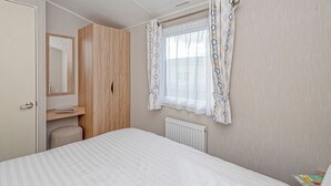 A bedroom with a bed, a wooden wardrobe, a window with patterned curtains, a radiator, and a small table with a magazine.