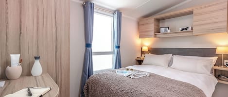 A hotel room with a bed, bedside tables, a desk, a window with curtains, and a wall-mounted shelf.
