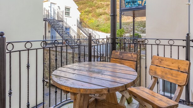 A wooden table and benches on a balcony with a metal railing, overlooking a building with stairs and a hillside.