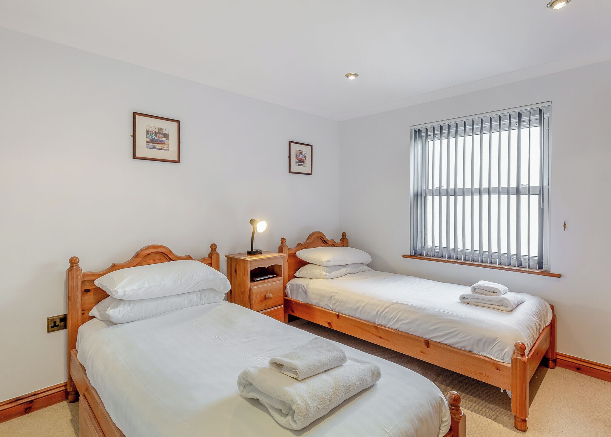 Une chambre à coucher avec deux lits en bois, une literie blanche, une table de chevet avec une lampe et une fenêtre avec des stores.