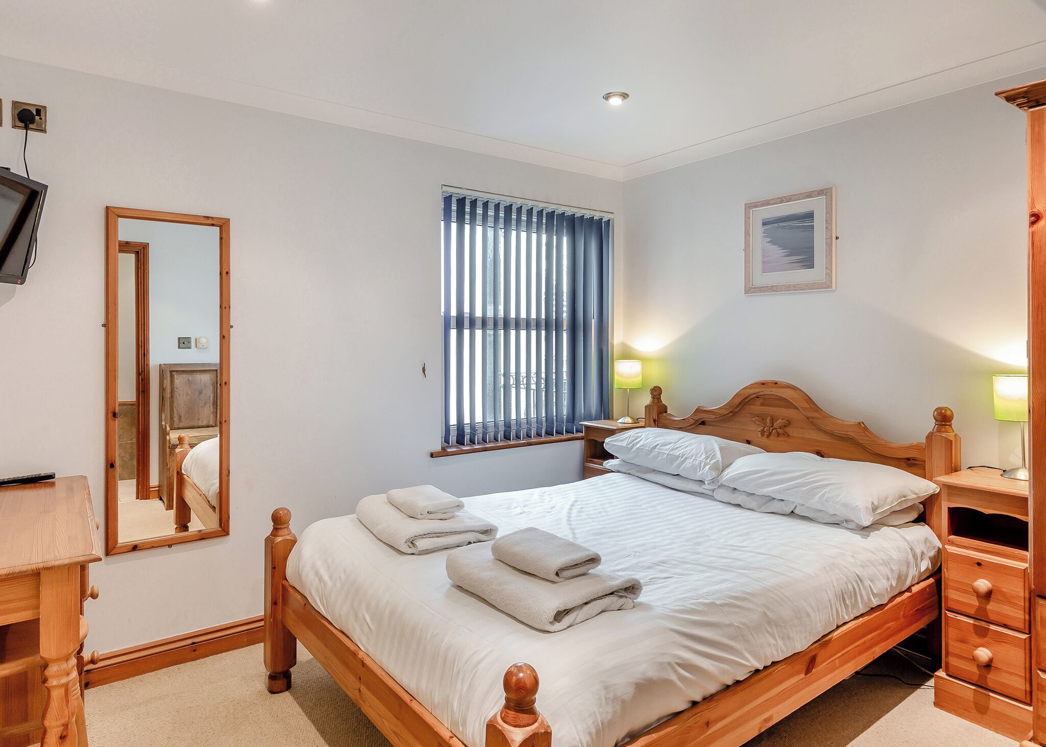 Quarto com cama de madeira, abajures, espelho, janela com persianas e televisão montada na parede.