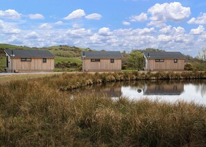 Three wooden cabins with sloped roofs, situated by a calm pond with tall grasses, under a partly cloudy sky.