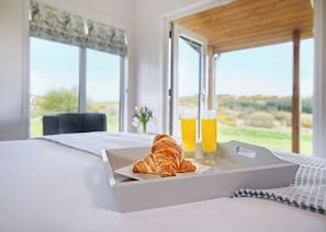 Un lit avec un plateau de petit-déjeuner contenant un croissant, deux verres de jus d’orange et un vase avec des fleurs.