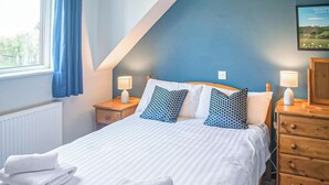 Un lit bien fait, avec du linge de lit blanc et des coussins à motifs bleus, une table de chevet en bois avec une lampe, et un mur bleu orné d’un tableau encadré.