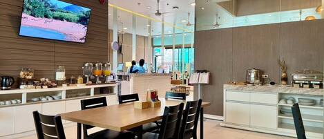 A modern dining area with a wooden table and chairs, a buffet counter with food and drinks, and a television displaying a scenic landscape.