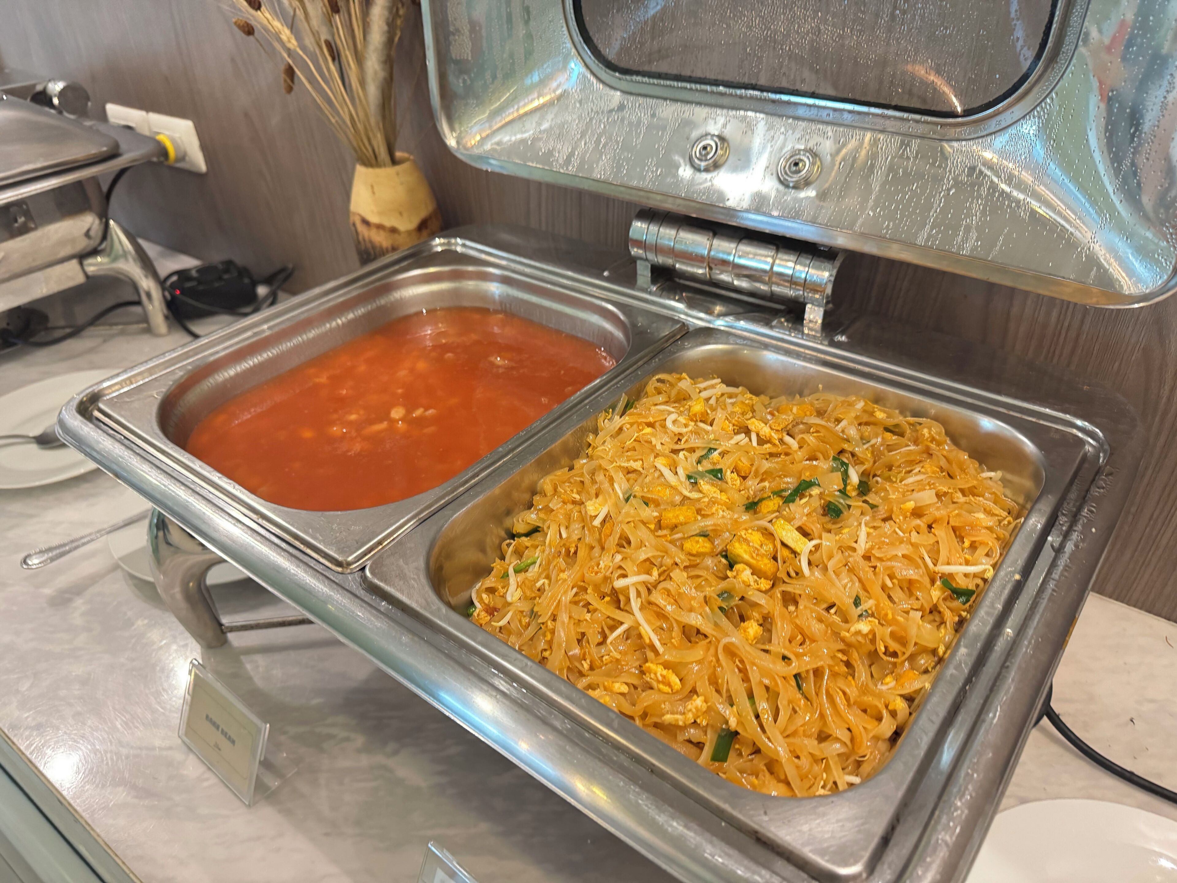 A buffet with two metal serving trays, one containing a red soup and the other noodles with eggs.