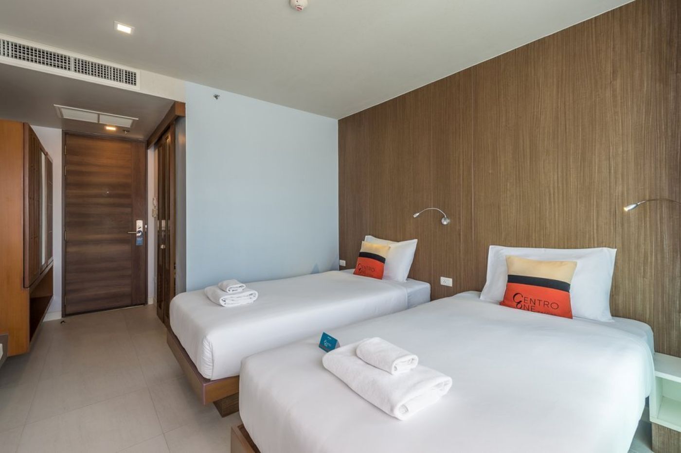 A hotel room with two beds, each with white bedding and pillows, a wooden accent wall, and a door leading to another room.