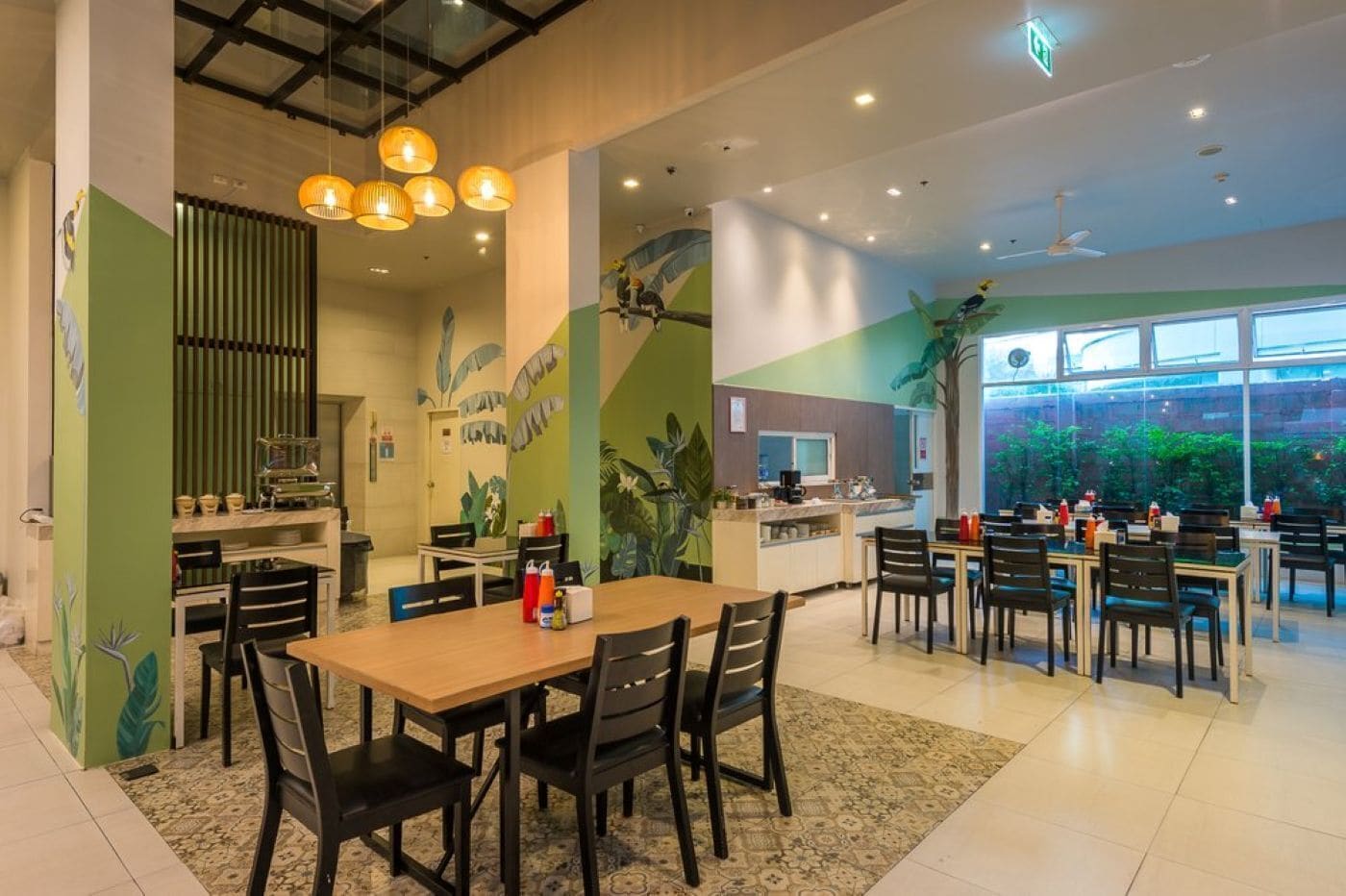A modern dining area with multiple tables and chairs, a kitchen area, and large windows with a view of greenery outside.