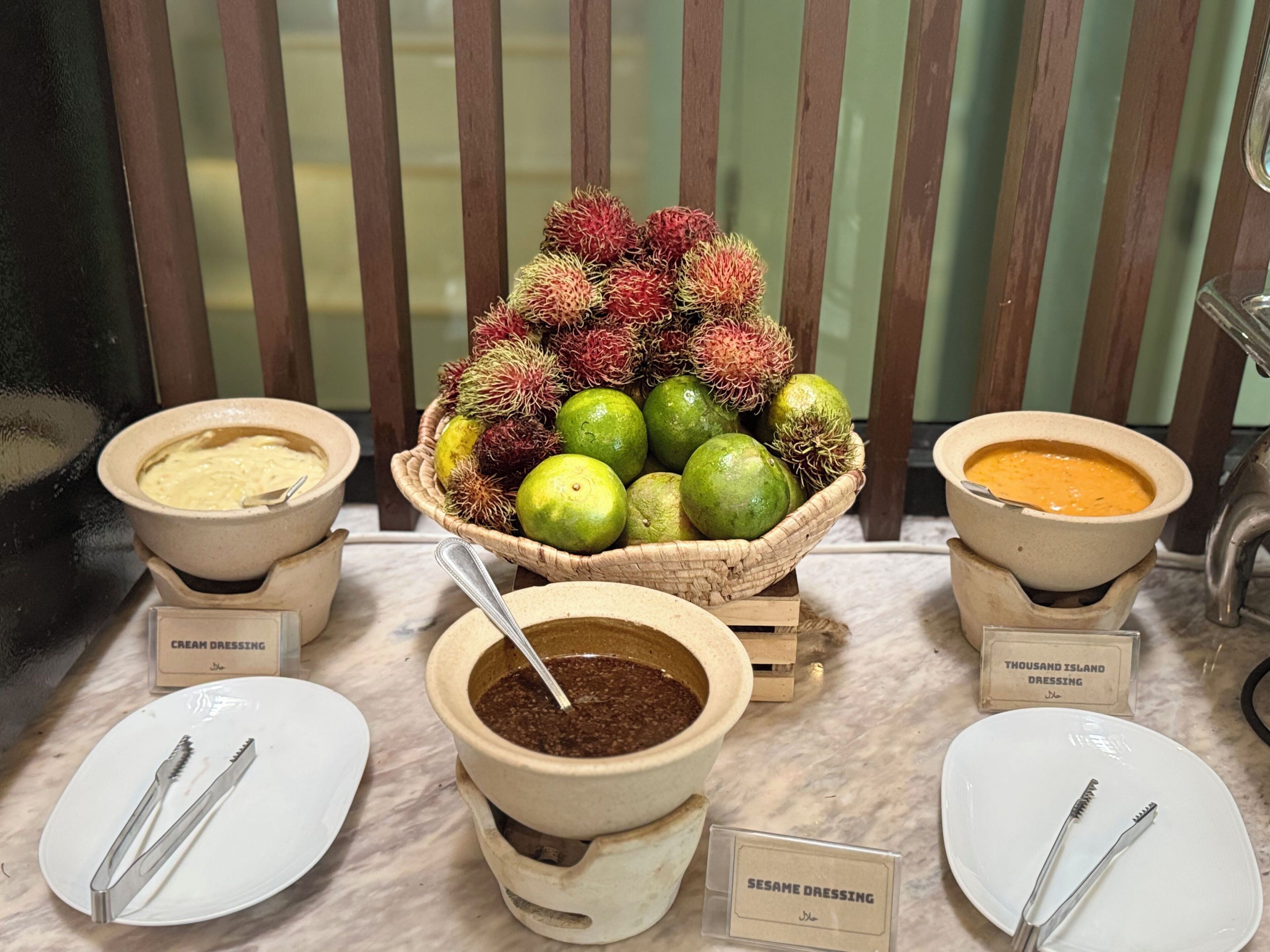 Ein Buffet mit beschrifteten Schalen für Sahndressing, Sesamdressing und einer zentralen Obstauslage mit Rambutan-Früchten und Limetten.