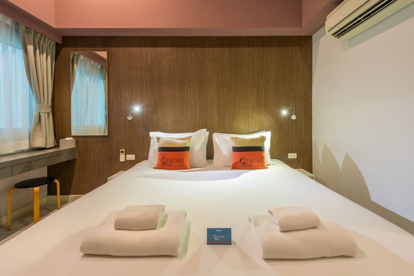 Une chambre d’hôtel avec une tête de lit en bois, deux oreillers portant la marque « CENTRO ONE », un lit avec du linge de lit blanc, une petite table avec une chaise et un climatiseur fixé au mur.