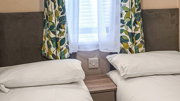 A hotel room with two single beds, a bedside table, a power outlet, and curtains with a leaf pattern.