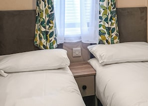 A hotel room with two single beds, a bedside table, a power outlet, and curtains with a leaf pattern.