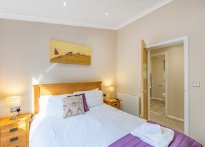 A hotel room with a bed, bedside lamps, a nightstand, a wooden headboard, a painting on the wall, and a bathroom door.