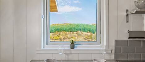 A kitchen with a window view of a green field, a sink, and a colander on the countertop.