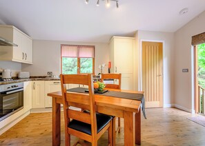 A modern kitchen with wooden cabinets, a dining table with chairs, a window with blinds, and a door leading to another room.