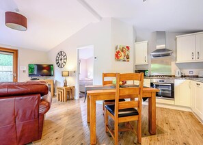 A modern living room with a leather sofa, a dining table with chairs, a TV, a clock, and a kitchen with appliances.