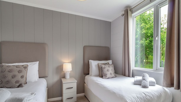 Un dormitorio con dos camas, una mesita de noche con lámpara y una ventana con cortinas.