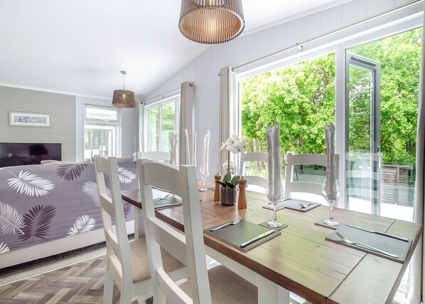 A dining area with a table set for two, white chairs, a vase with flowers, and a view of greenery through large windows.