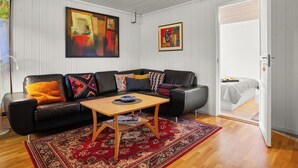 A living room with a black leather sofa, a wooden coffee table, and a red patterned rug. There are two paintings on the wall, and a door leading to a bedroom. - Krokenskogen (Sandefjord)