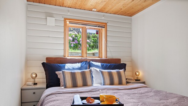 A neatly made bed with a tray of breakfast items, a bedside lamp, and a window with a view of flowers. - Krokenskogen (Sandefjord)
