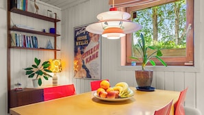 A dining area with a wooden table, red chairs, a fruit bowl, a potted plant, a poster, and a window with a view of trees. - Krokenskogen (Sandefjord)