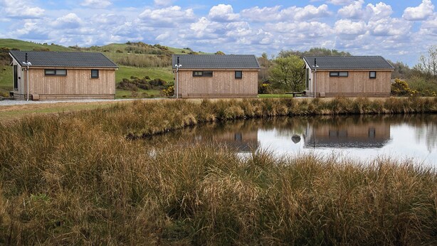 Three wooden cabins with sloped roofs, situated on a grassy hill with a calm pond in the foreground. - This Studio Lodge is perfect for couples. (Ringford)