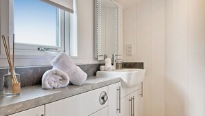 A modern bathroom with a large mirror, white cabinets, a marble countertop, and a window with blinds. - This Studio Lodge is perfect for couples. (Ringford)
