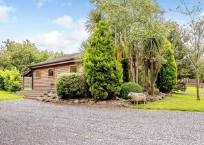 A log cabin with a gravel driveway, surrounded by various trees and a small sheep statue.