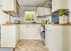 A compact kitchen with white cabinetry, a herringbone-patterned floor, a window with blinds, and a hanging plant.