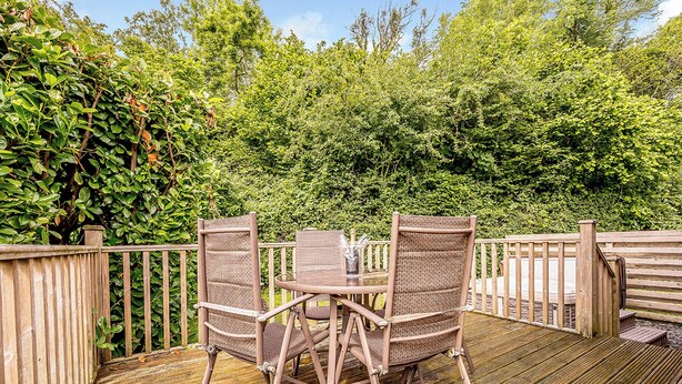 A wooden deck with two wicker chairs and a glass table, surrounded by dense greenery. - Deluxe lodge with one master double with en suite walk-in shower and one twin. (Cartmel)