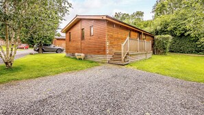 A wooden cabin with a porch, gravel driveway, and a red car parked nearby. - Deluxe lodge with one master double with en suite walk-in shower and one twin. (Cartmel)