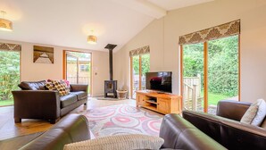 A spacious living room with a large TV, leather sofas, a fireplace, and large windows offering a view of greenery. - Deluxe lodge with one master double with en suite walk-in shower and one twin. (Cartmel)