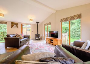 A spacious living room with a large TV, leather sofas, a fireplace, and large windows offering a view of greenery.