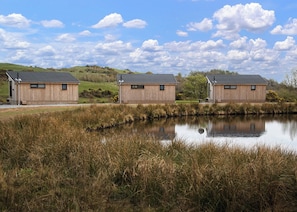 Trois cabanes en bois aux toits inclinés, situées au bord d’un étang paisible bordé de hautes herbes, sous un ciel partiellement nuageux.