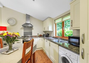 A kitchen with a stove, microwave, and washing machine, a clock on the wall, and a window with a view of greenery.