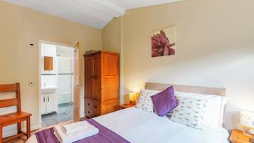 A bedroom with a bed, a wooden wardrobe, a nightstand, a chair, and a bathroom visible through an open door.