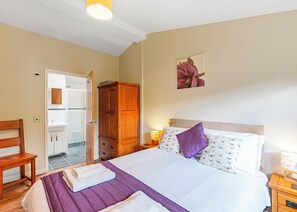 A bedroom with a bed, a wooden wardrobe, a nightstand, a chair, and a bathroom visible through an open door.